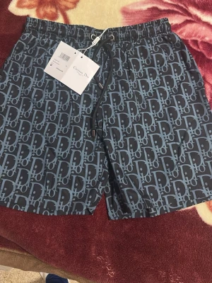 Blå badbyxor från Christian Dior - Snygga badbyxor från Christian Dior i blått med all-over logomönster. De har elastisk midja med dragsko och innerbyxa i mesh för extra komfort. Perfekta för stranden eller poolen och ger en lyxig känsla till din sommarstil.