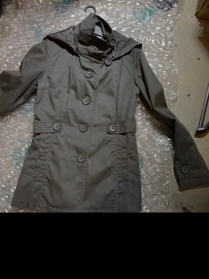 Olivgrön trenchcoat med huva - Stilren olivgrön trenchcoat med dubbelknäppning och stora knappar. Jackan har en praktisk huva, markerad midja med dekorativa knappar och långa ärmar med knappdetaljer vid ärmslut. Tillverkad i ett slätt tyg som ger en klassisk look.