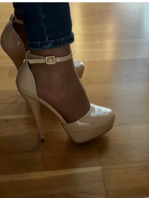 Beige platåpumps med ankelrem - Snygga beige pumps i lack med hög platå och smal klack. Skorna har rund tå och justerbar ankelrem med guldfärgat spänne. Perfekta för dig som vill ha extra höjd och en elegant look.