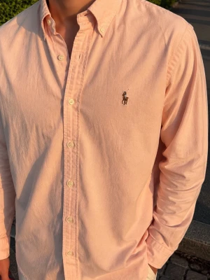 Ljusrosa skjorta från Ralph Lauren - Klassisk ljusrosa skjorta från Ralph Lauren med button-down krage och broderad logga på bröstet. Skjortan har långa ärmar, normal passform och är tillverkad i mjuk bomull. Perfekt för en clean och stilren look.