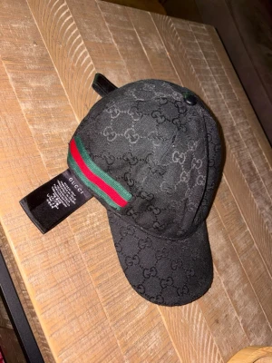 Svart keps från Gucci med logomönster - Snygg svart keps från Gucci med diskret GG-logomönster över hela kepsen. På sidan finns den klassiska Gucci-randen i rött och grönt. Kepsen har böjd skärm och justerbar passform. Perfekt för dig som vill ha en stilren accessoar med lyxig känsla.