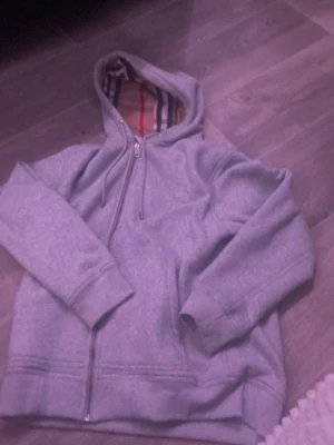 Grå hoodie från Burberry med dragkedja - Säljer en grå hoodie från Burberry med dragkedja och klassiskt rutigt foder i huvan. Hoodien har broderad logga på bröstet, ribbade muddar och är gjord i mjukt bomullsmaterial. Perfekt för dig som gillar stilrena och exklusiva plagg.