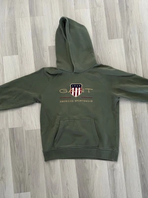 Olivgrön hoodie från GANT - Snygg olivgrön hoodie från GANT med stor huva och känguruficka. På bröstet finns ett broderat GANT-emblem med texten '1949 American Sportswear' och rödvita detaljer. Hoodien har en avslappnad passform och är tillverkad i mjukt bomullsmaterial.