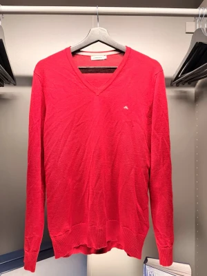  J.Lindeberg - Snygg röd V-ringad tröja från J.Lindeberg med diskret logga på bröstet. Tröjan har långa ärmar och ribbade muddar vid ärmslut och nederkant. Perfekt för dig som gillar stilrena plagg med en poppig färg.
