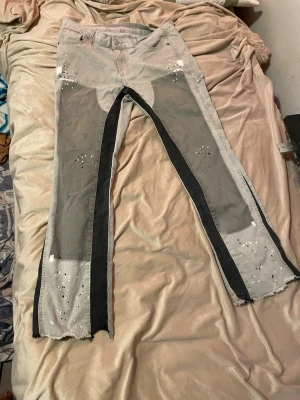 Grå jeans med svarta detaljer - Unika grå jeans med svarta paneler längs benen och råa, fransiga benslut. Jeansen har vita färgstänk och små håldetaljer för en edgy look. Modellen är rak och något utsvängd, med flera fickor och patchdetaljer på baksidan.