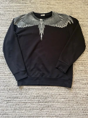 Svart sweatshirt med vingtryck från Marcelo Burlon - Säljer en svart sweatshirt från Marcelo Burlon med ikoniskt vingtryck i grått och vitt över axlar och bröst, samt på ryggen. Tröjan har rund halsringning, långa ärmar och är gjord i mjukt bomullsmaterial. Perfekt för dig som vill sticka ut med en unik design. 