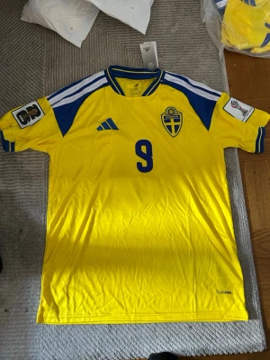 Sverige matchtröja Adidas gul/blå - Säljer en officiell svensk landslagströja från Adidas i gult med blå detaljer och nummer 9 på bröstet. Tröjan har Sveriges landslagsemblem, Climacool-material och tryck på ärmarna. Perfekt för fotbollsfans som vill stötta Sverige på matchdag!