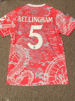 Real Madrid Bellingham matchtröja rosa - Säljer en rosa Real Madrid matchtröja från Adidas med Bellingham och nummer 5 på ryggen. Tröjan har ett unikt drakmönster i vitt och grått, klubbmärke på bröstet och sponsortryck framtill. Korta ärmar och rund hals. Perfekt för fotbollsfans!