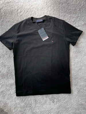 Svart t-shirt från Tommy Hilfiger - Klassisk svart t-shirt från Tommy Hilfiger med rund halsringning och diskret logga broderad på bröstet. Tillverkad i mjuk bomull för en bekväm känsla. Perfekt basplagg till garderoben.