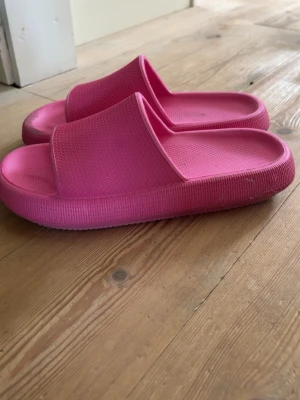 Rosa tofflor - Säljer ett par chunky rosa tofflor i mjukt och flexibelt gummimaterial. De har en bred rem över foten och en tjock sula som ger skön dämpning. Perfekta att glida runt i hemma, ute eller på stranden. Färgen är riktigt poppig och ger en cool vibe. De är från skopunkten men denna färg säljs troligen inte längre. Storlek 41, använda 2-3 gånger.