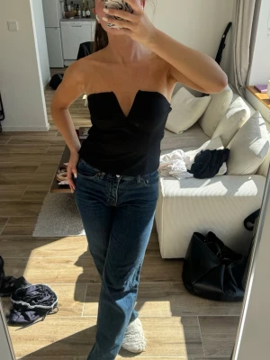 Svart axelbandslös tubtopp - Snygg svart axelbandslös tubtopp med en djup v-ringning framtill. Toppen har en croppad passform och är perfekt för att styla med jeans eller kjol. Materialet känns mjukt och följsamt mot kroppen.