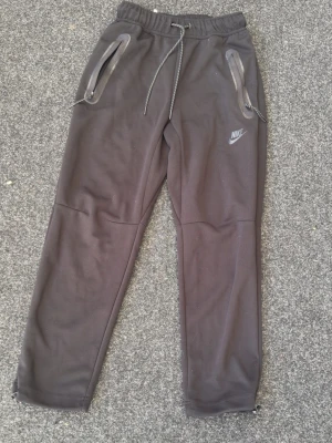 Svarta mjukisbyxor från Nike - Snygga svarta mjukisbyxor från Nike med dragsko i midjan och två dragkedjeförsedda fickor. Byxorna har en avslappnad passform och Nike-logga på vänster ben. Perfekta för chill eller träning.