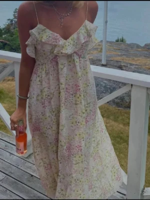 Blommig långklänning med volang - Romantisk långklänning med tunna axelband och volang vid bysten. Klänningen har ett blommigt mönster i ljusa toner av rosa, grön och gul på vit botten. Perfekt för varma sommardagar och har en luftig passform.