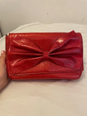 Röd glittrig kuvertväska med rosett - Snygg röd kuvertväska med glittrigt material och stor dekorativ rosett på framsidan. Väskan har en guldfärgad kedja som axelrem och en rektangulär form. Perfekt för att ge din outfit extra bling.