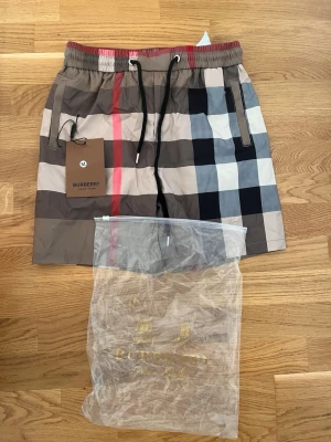 Rutig Burberry shorts med snörning - Snygga shorts från Burberry med klassiskt rutigt mönster i beige, svart, vitt och rött. Shortsen har elastisk midja med snörning och fickor på sidorna samt en bakficka. Perfekta för sommaren och ger en trendig look.