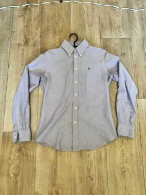 Ljuslila  skjorta från Ralph Lauren - Klassisk ljuslila skjorta från Ralph Lauren med button-down krage och broderad logga på bröstet. Skjortan har långa ärmar, vita knappar och är tillverkad i mjuk bomull. Perfekt för en clean och stilren look. Pris går alltid att diskutera. Sjukt bra plagg till sommaren att slänga på under en väst eller använda som den är 