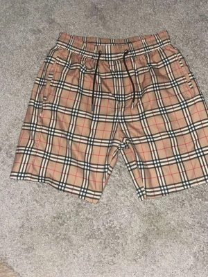 Beige rutiga shorts med snörning - Säljer ett par beige shorts med klassiskt rutigt mönster i svart, vitt och rött. Shortsen har elastisk midja med svart snörning och sidofickor. Perfekta för sommaren och riktigt snygga till en enkel t-shirt.