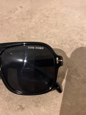 Svarta solglasögon från Tom Ford - Snygga svarta solglasögon från Tom Ford med klassisk pilotform och mörka glas. Bågarna är i blank svart plast med gulddetaljer vid gångjärnen och en unik bro ovanför näsan. Perfekta för dig som vill ha en stilren och trendig look.