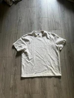 Vit ribbad t-shirt från Bershka - Säljer en stilren vit t-shirt från Bershka i ribbad bomull. T-shirten har korta ärmar, rund halsringning och en relaxed passform. Perfekt basplagg att matcha med allt i garderoben.