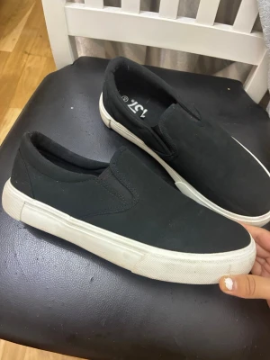 Lager 157 Slip-on skor i svart, storlek 40 - Ett par slip-on skor i svart med vit sula. Skorna har resår på sidorna för enkel på- och avtagning. Helt nya. Storlek 40. 