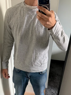 Ljusgrå sweatshirt med raglanärm - En stilren ljusgrå sweatshirt med raglanärmar och rund hals. Tröjan är i mjukt bomullsmaterial och har en klassisk V-detalj vid halsringningen. Perfekt för en avslappnad look och enkel att matcha med jeans eller joggers.