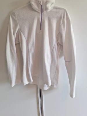 Vit fleece-tröja med halv dragkedja - Mysig vit fleece-tröja med lång ärm och halv dragkedja framtill. Tröjan har en hög krage och diskreta sömmar i ljuslila. Perfekt för lager-på-lager