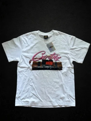 Vit t-shirt med tryck och grafiskt motiv - Vit t-shirt med ett stort grafiskt tryck framtill som föreställer texten 'Crazy' i rosa och en digital timer. Klassisk passform och rund halsringning. Perfekt för dig som gillar unika och uttrycksfulla plagg.