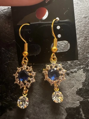 Guldiga örhängen med blå sten - Snygga örhängen i guldton med gnistrande blå mittsten omgiven av små klara stenar i en stjärnformad design. Under hänger en rund klar sten som ger extra bling. Perfekta för dig som gillar glam och detaljerade smycken.