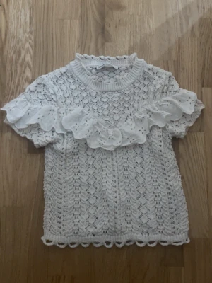 Stickat Vit spetsblus med volang - Supersöt vit blus med spetsmönster och en bred volang över bröstet och ärmarna. Kortärmad modell med rund hals och fina virkade detaljer längs kanterna. Perfekt för dig som gillar romantisk och feminin stil.