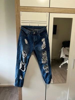Blå ripped mom jeans - Säljer ett par blå mom jeans med kraftiga slitningar och hål framtill. Jeansen har hög midja, raka ben och klassisk femficksdesign. Perfekta för en avslappnad och trendig streetstyle-look.