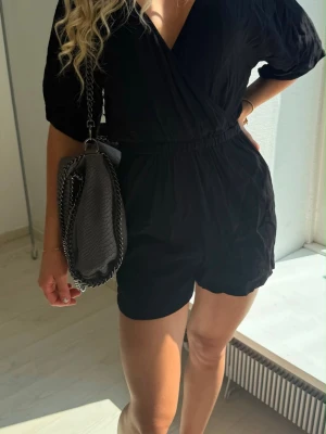 Svart kort jumpsuit med omlott - Snygg svart jumpsuit med omlott-ringning och resår i midjan. Modellen har korta ben och halvlånga ärmar, vilket ger en avslappnad men trendig look. Perfekt till sneakers eller boots. Materialet ser ut att vara mjukt och följsamt, troligen bomull eller viskos.
