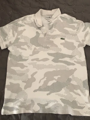 Lacoste pike  - Säljer en vit henley från Lacoste med grått camouflage-mönster och klassisk krage. Skjortan har korta ärmar och den ikoniska gröna krokodilloggan på bröstet. Perfekt för dig som gillar streetwear och vill sticka ut lite extra.