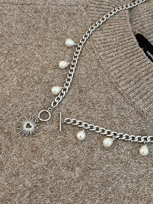 Silverfärgat halsband med pärlor - Snyggt halsband i silverfärgad rostfritt med kedja och vita pärlor som hänger längs hela halsbandet. Halsbandet har även ett solformat hänge med ett hjärta i mitten. Perfekt accessoar för att lyfta din outfit.