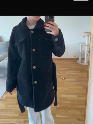 Svart teddyjacka med bälte - Mysig svart teddyjacka med stora knappar framtill och bälte i midjan. Jackan har två bröstfickor och en avslappnad passform. Perfekt för kalla dagar när du vill vara både varm och snygg. Hål i en av sidofickorna