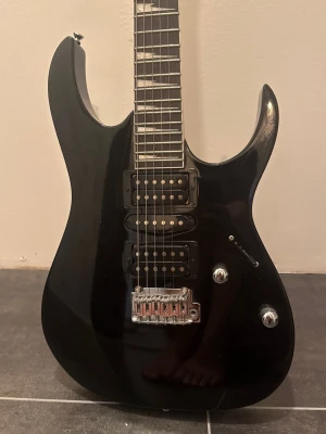Ibanez GIO - Model: GRG170DX-BKN .Säljer en svart elgitarr med två humbucker-mickar och en single coil i mitten. Kroppen har en modern, vass form och glänsande finish. Gitarren har två rattar och en switch för att justera ljudet samt en metallisk stall. Perfekt för dig som gillar rock eller metal.
