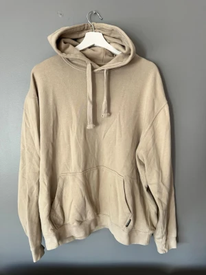 Beige oversized hoodie med huva - En stilren beige hoodie med huva och dragsko, samt en stor känguruficka framtill. Hoodien har en avslappnad, oversized passform och är tillverkad i mjukt bomullsmaterial. Perfekt för en chill och trendig look.