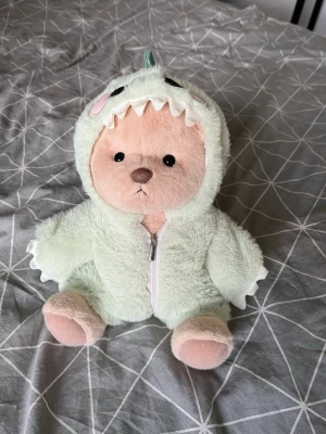 Söt nalle i mintgrön dinosauriedräkt  - Mjuk och gullig nallebjörn med ljusbrun päls, klädd i en mintgrön plyschdräkt med dragkedja och dinosauriedetaljer som taggar och tänder på huvan. Från Get A Hug