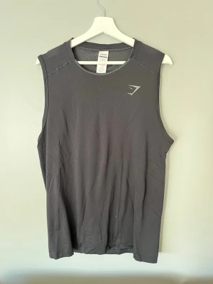 Svart träningslinne från Gymshark - Säljer ett svart träningslinne från Gymshark i storlek L. Linnet är tillverkat i ett lätt och ventilerande polyestermaterial, perfekt för gymmet eller löprundan. Ärmlös design med diskret Gymshark-logga på bröstet. Snygg och funktionell för alla träningspass.