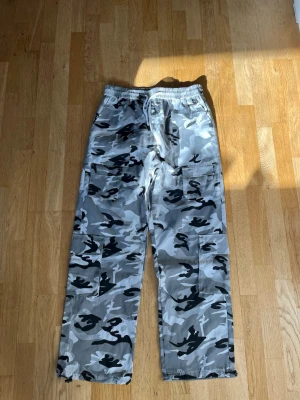 Grå camo cargobyxor  - Säljer ett par cargobyxor i grått camomönster med elastisk midja och vit dragsko. Byxorna har stora sidofickor och raka ben. Perfekta för en avslappnad streetwear-look. Materialet känns lätt och bekvämt.