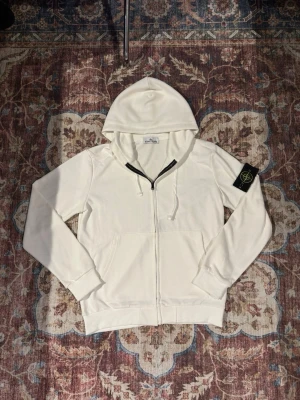 Vit hoodie från Stone Island - Säljer en stilren vit hoodie från Stone Island med dragkedja framtill och klassisk huva. Hoodien har en svart logotyp-patch på vänster ärm och fickor på magen. Tillverkad i mjukt bomullsmaterial och har ribbade muddar vid ärmslut och nederkant.