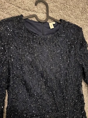 Svart glittrig långärmad klänning - Snygg svart klänning med långa ärmar och massor av glittriga paljetter. Perfekt för dig som vill sticka ut med en elegant look som väl oxh bröllop. Klänningen har rund halsringning och figurnära passform.