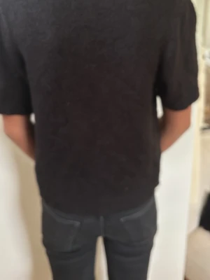 Svart kortärmad kofta med knappar - Kortärmad svart kofta med v-ringning och fyra dekorativa knappar framtill. Koftan har ribbade muddar vid ärmslut och nederkant, och är gjord i ett mjukt, stickat material. Perfekt att slänga över ett linne för en chill look.