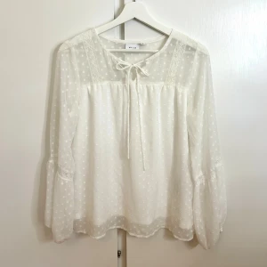 Vit blus från Vila - Supersöt vit blus från Vila med transparenta, prickiga ärmar och spetsdetaljer. Blusen har knytband vid halsen och ballongärmar som ger en romantisk vibe. Perfekt till ljusa jeans eller kostymbyxor för en fräsch look.