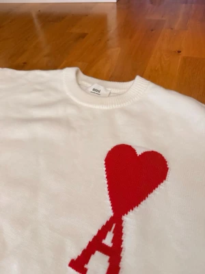 Vit stickad tröja från Ami Paris - Vit stickad tröja från Ami Paris med röd hjärtlogga och bokstaven A på bröstet. Tröjan har rund halsringning, långa ärmar och ribbade muddar vid ärmslut och nederkant. Perfekt för dig som gillar stilrena plagg med en twist.