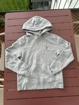 Grå hoodie från Polo Ralph Lauren - Snygg grå hoodie från Polo Ralph Lauren med klassisk huva och känguruficka framtill. Tröjan har det ikoniska lilla blå logobroderiet på bröstet och är tillverkad i mjukt bomullsmaterial. Perfekt för en avslappnad och stilren look.
