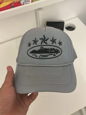 Ljusgrå truckerkeps med stjärnor - Säljer en ljusgrå truckerkeps med svart broderad design framtill, inklusive fem stjärnor och ett ovalt motiv. Kepsen har mesh på sidorna och bak för extra ventilation samt fler broderade stjärnor på sidan. Justerbar baktill för perfekt passform.