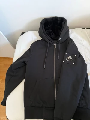 Svart pufferjacka från Moose Knuckles - Säljer en svart pufferjacka från Moose Knuckles med huva och dragkedja framtill. Jackan har ett emblem på ärmen, två fickor och mjukt foder på insidan. Perfekt för kalla dagar och riktigt skön att bära.