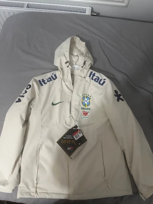 Vit Brasil Nike vindjacka med huva - !!!Står storlek L men är passar som S/M!!!Snygg vit vindjacka från Nike med Brasil-logga och sponsormärken. Jackan har huva med dragsko, dragkedjefickor och mjukt grått innerfoder. Perfekt för dig som gillar sportig stil och vill sticka ut med landslagskänsla. En riktig Gore-Tex vara.