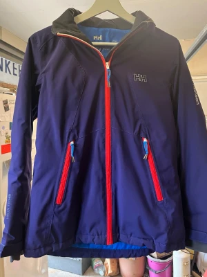 Blå Helly Hansen vindjacka med huva - Snygg mörkblå vindjacka från Helly Hansen med röd dragkedja och blå detaljer. Jackan har huva med ljusblå insida, två framfickor med dragkedja och Helly Tech-material som står emot vind och vatten. Perfekt för blåsiga dagar och outdoor-äventyr. Armhåla till armhåla ca: 45cm. Axel till midjan ca: 60cm. Armlängd ca: 45cm. Midjan ca: 50cm