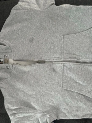 Grå hoodie från Burberry med dragkedja - Snygg grå hoodie från Burberry med dragkedja framtill och klassiskt rutigt foder i huvan. Hoodien har fickor på magen och diskret Burberry-logga på bröstet. Perfekt för en avslappnad men stilren look.
